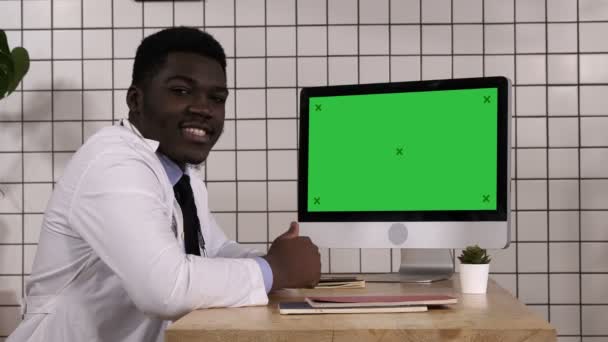 Black doctor talking to camera sitting next to computer screen. White Display. — Stock Video