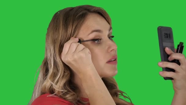 Gyönyörű szőke fiatal nőt alkalmazó nézett tükörbe smartphone egy zöld képernyő, Chroma Key szempillaspirál. — Stock videók
