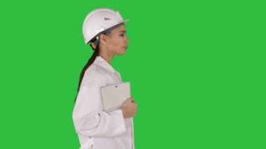Holding dizüstü veya tablet yeşil ekranda Chroma Key yürüyüş Beyaz laboratuar ceket ve beyaz Emanet baret çekici İspanyol kadın.
