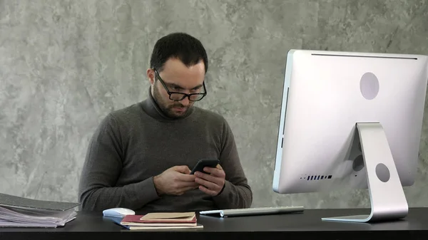 Sakallı genç işadamı modern ofiste çalışıyor. Onun smartphone içinde seyir ve bir şey yazarak adam. — Stok fotoğraf