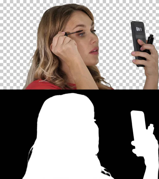 Gyönyörű szőke fiatal nő alkalmazó szempillaspirál néz smartphone tükör, Alpha Channel — Stock Fotó