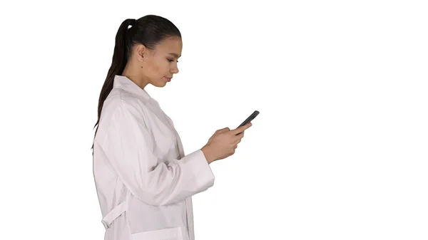 Alegre linda mujer joven médico escribiendo texto en el teléfono mientras camina sobre fondo blanco. —  Fotos de Stock