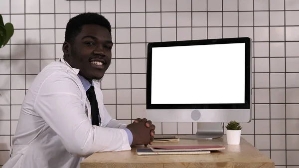 Médico negro falando com câmera sentada ao lado da tela do computador. Exposição branca. — Fotografia de Stock