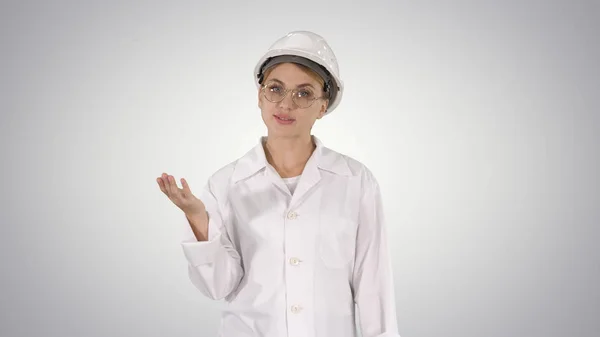 Mujer ingeniera madura con sombrero duro y bata de laboratorio hablando y presentando algo apuntando a los lados en el fondo del gradiente. —  Fotos de Stock