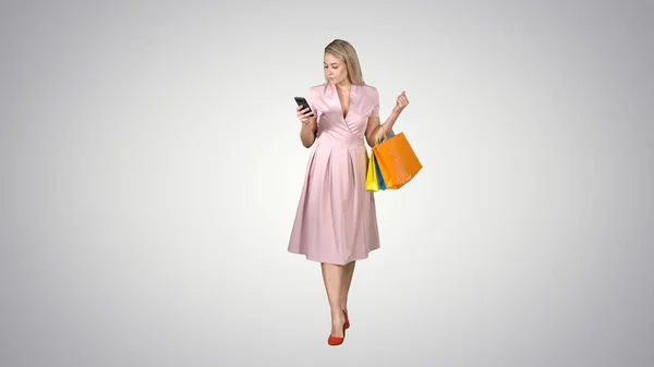 Shopping woman with bags texting message on smartphone while walking on gradient background. — Stock Photo, Image