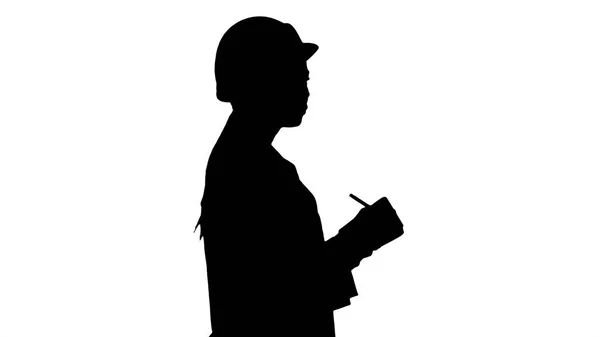 Sylwetka Kobieta inżynierem z kask trzyma długopis i Lista kontrolna wprowadzenie coś w dół podczas chodzenia. — Zdjęcie stockowe