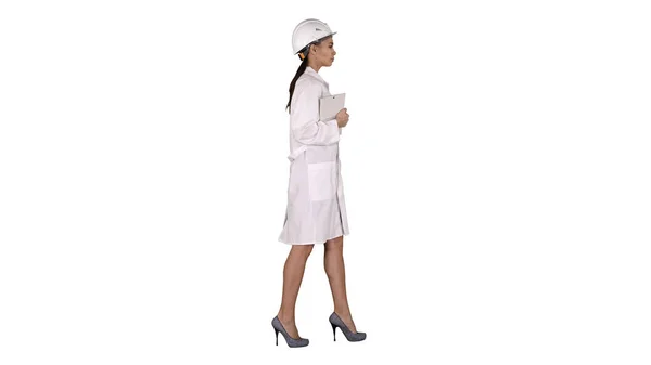 Atractiva mujer hispana con bata blanca de laboratorio y sombrero blanco de seguridad caminando sosteniendo cuaderno o tableta sobre fondo blanco . —  Fotos de Stock