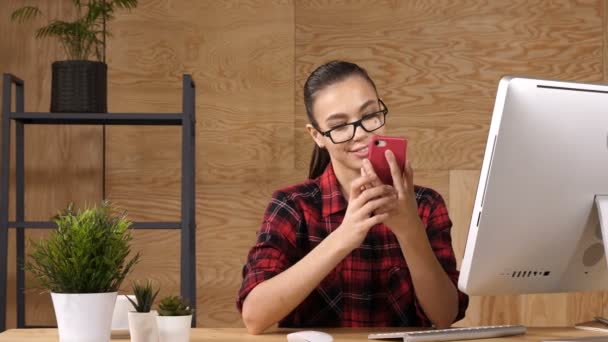 Jeune femme moderne envoyant un message texte sur son téléphone intelligent assis près de l'ordinateur . — Video