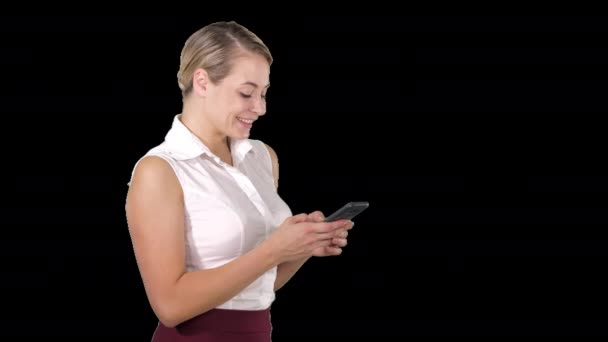 SMS à un ami Jolie jeune femme tenant un téléphone intelligent et le regardant avec le sourire tout en se tenant debout, Alpha Channel — Video