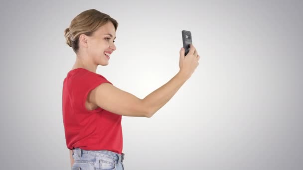 Beautiful young woman walking and holding a smartphone up to take pictures and selfies on gradient background. — Stock Video