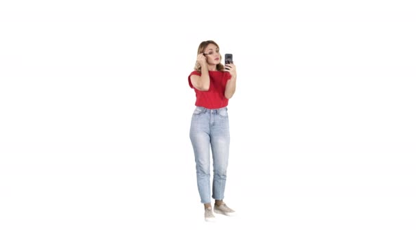 Mulher bonita usando rímel e olhando em seu smartphone no fundo branco. — Vídeo de Stock