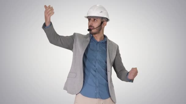 Architecte homme dans le casque et la tête mis parler à la caméra et faire des gestes montrant sur certains objets sur fond de dégradé. — Video