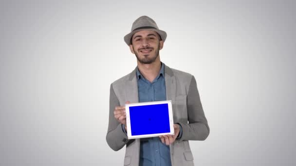 Hombre casual en un sombrero caminando y mostrando la tableta digital a la cámara en el fondo de gradiente. — Vídeo de stock