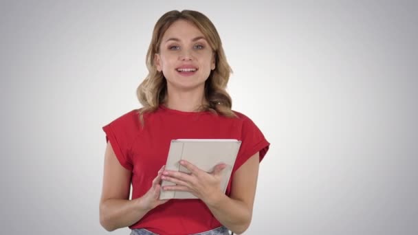 Kobieta prezenterka w czerwonym t-shirt trzyma tablet obracając strony i rozmawia z kamerą na tle gradientu. — Wideo stockowe