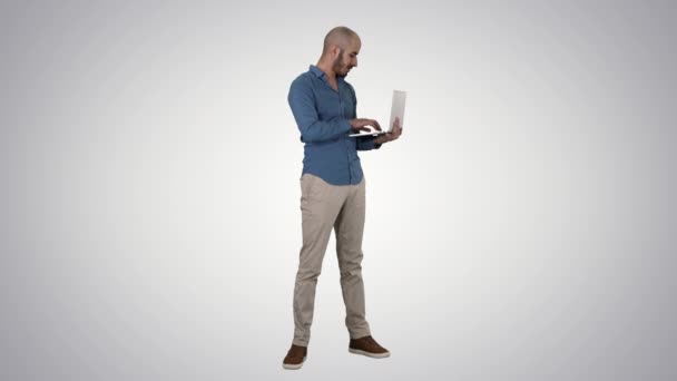 Unga Mellanöstern affärsman, stående och skriva på laptop när du håller den i händerna på gradient bakgrund. — Stockvideo
