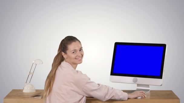 Glad kvinna sitter vid bordet med en dator i Office och tittar på kamera leende blå skärm mock-up display på gradient bakgrund. — Stockvideo