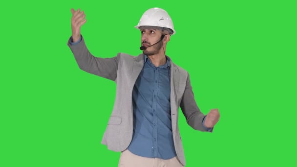 Arquitecto hombre en casco y conjunto de cabeza hablando con la cámara y haciendo gestos que se muestran en algunos objetos en una pantalla verde, Chroma Key . — Vídeos de Stock