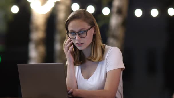 Atractiva joven mujer que tiene una conversación de teléfono inteligente mientras trabaja en un ordenador portátil . — Vídeos de Stock