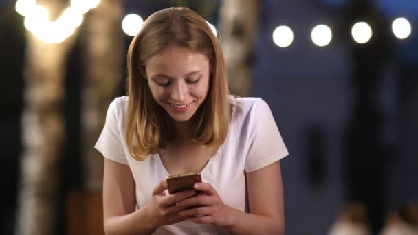 Genç ve çekici bir kız yazın akşam akıllı telefonuna mesaj yazıyor. Gülümsüyor, iletişim kuruyor.. — Stok video