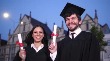 Diplomalarını gösteren iki mutlu mezun öğrenci kameraya poz veriyor. Rüzgar kızların kafalarını uçuruyor..