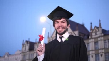 Mezuniyet kıyafeti adam gülümseyerek ve kameraya diploma gösteren.