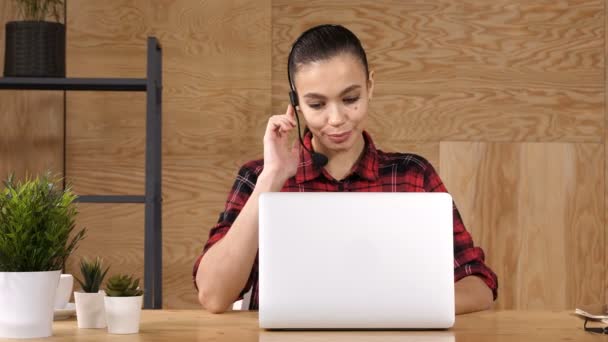 Freelancer bonita fêmea posando e trabalhando on-line com fone de ouvido com microfone em um pequeno desktop de escritório ou em casa . — Vídeo de Stock