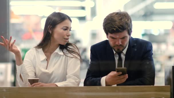 Hombre de negocios trabajando en un teléfono móvil mientras frustrado chica aburrida discutiendo sentado a su lado en el café del centro comercial . — Vídeo de stock