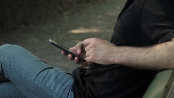 Mannelijke handen met smartphone in een park. Zittend op een bank. — Stockvideo