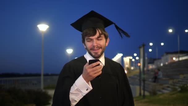 Mladý usměvavý muž absolvent drží smartphone textové něco a chůzi. — Stock video