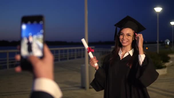 Feliz estudiante graduada sosteniendo un diploma posando mientras su amiga le toma una foto con su smartphone. — Vídeo de stock