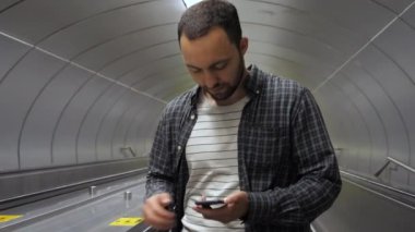 Pozitif neşeli hipster adam yürüyen merdivende dururken telefonla mesaj atıyor..
