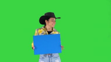 Gülümseyen güzel cowgirl boş tahta tutarak ve yeşil ekran üzerinde dans, mavi mockup.