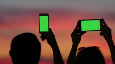 Bir adam ve bir kadın telefonlarında gün batımının fotoğraflarını çekiyorlar, onları arkadan görüyoruz..