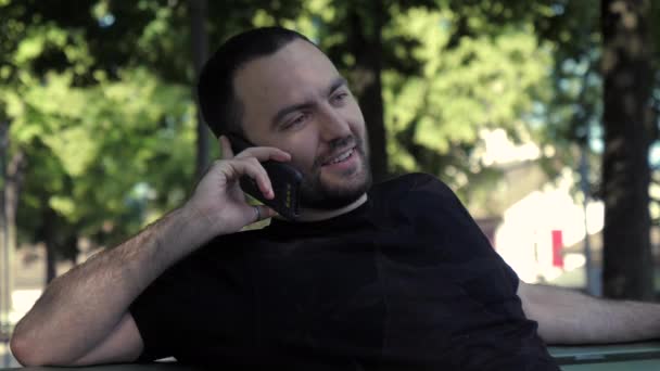 Un joven guapo sentado en un banco hablando por teléfono en el parque. — Vídeo de stock