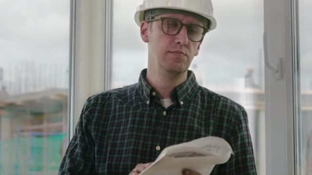 Bauingenieur trägt Sicherheitshelm Blick auf Blaupause Papier Konstruktionsplan auf dem Hintergrund des Fensterrahmens mit blau gelb und Baustelle. — Stockvideo