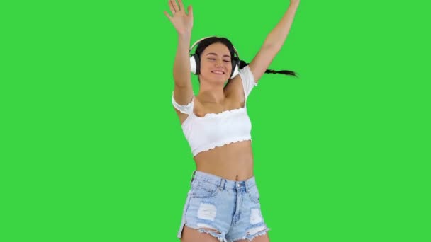 Vit kvinnlig modell i hörlurar hoppar, uttrycker glada känslor lyssna på musik på en grön skärm, Chroma Key. — Stockvideo