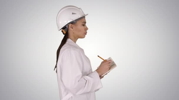 Ingeniera con casco sostiene bolígrafo y lista de verificación poniendo algo abajo mientras camina sobre fondo degradado. — Vídeos de Stock