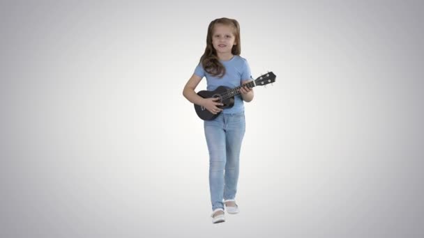 Liten flicka sjunger och spelar liten gitarr när man går mot kameran på gradient bakgrund. — Stockvideo