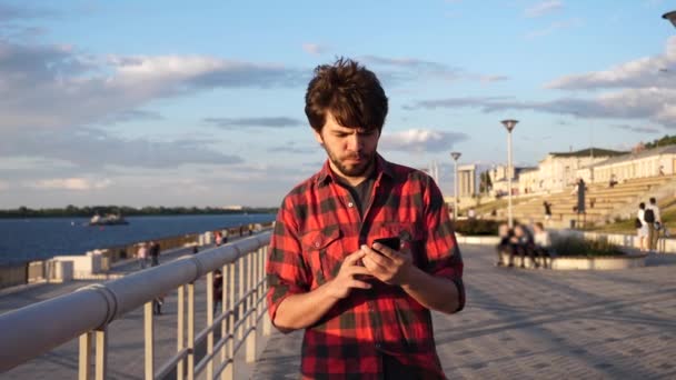 Homme décontracté marchant à l'extérieur lisant un message texte sur son téléphone intelligent. — Video