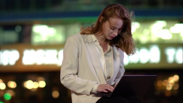 Una mujer de negocios sentada frente a un edificio de negocios y escribiendo en un portátil . — Vídeo de stock