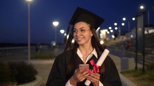Chica graduada de la escuela secundaria después de la caremonía de graduación caminando y mensajes de texto en el teléfono móvil. — Vídeos de Stock
