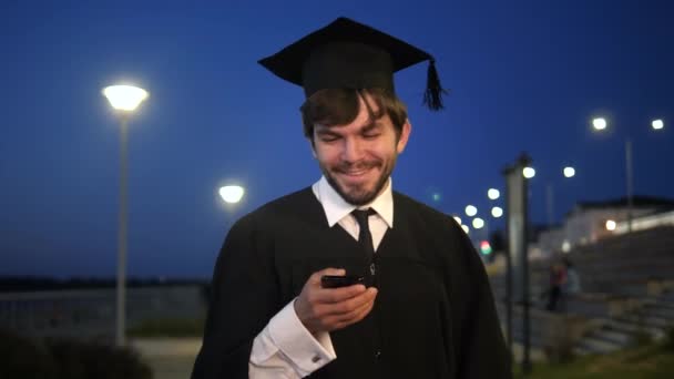 Estudiante graduado caminando por la noche y mensaje de texto en el teléfono . — Vídeos de Stock