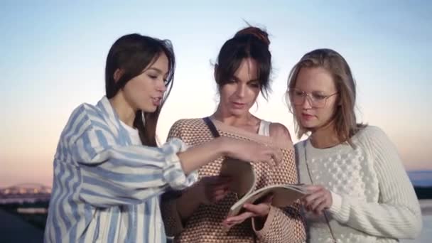 Three women friends outside looking through magazine. — Stock Video