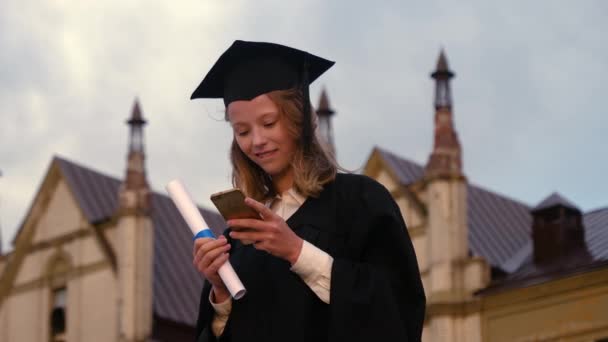 Söt tonåring i examensdräkt sms:ande på mobiltelefon. — Stockvideo