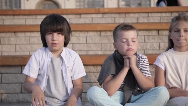 Cuatro niños que parecen aburridos y cansados sentados en un banco . — Vídeo de stock