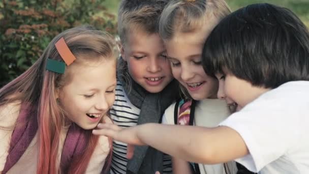 Cuatro niños felices se sientan al aire libre y usan el teléfono móvil sonriendo . — Vídeo de stock