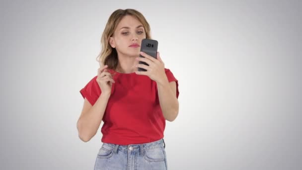 Une jolie jeune femme redresse ses cheveux en regardant dans le mobile sur fond de dégradé . — Video