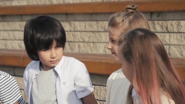 Verschiedene Kindergruppen sitzen gemeinsam auf einer Bank und warten. — Stockvideo