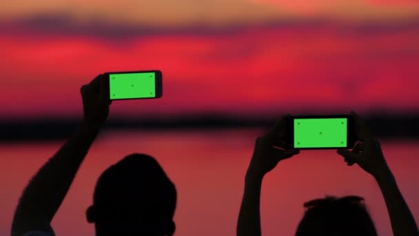 Silueta de un hombre y una mujer grabando vídeos de la puesta del sol con sus smartphones . — Vídeos de Stock