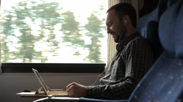 Casual businessman in train with tablet and headphones watching video and smiling. — Stock Video